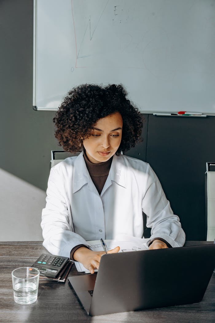 African American woman in office setting focused on work at laptop, with curly hair and business attire.
